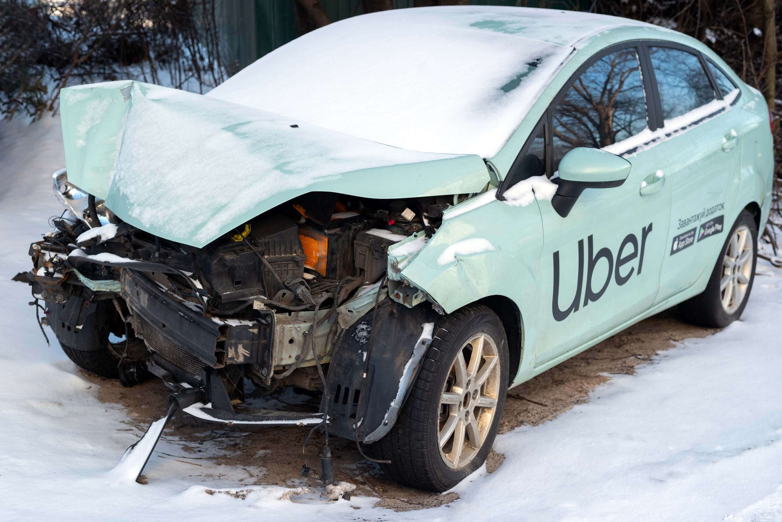 A light-colored sedan with a prominent Uber logo on the side door sitting in the snow with severe front-end crash damage, demonstrating a fatal collision scenario that leads to wrongful death after an Uber or Lyft accident in Oklahoma.
