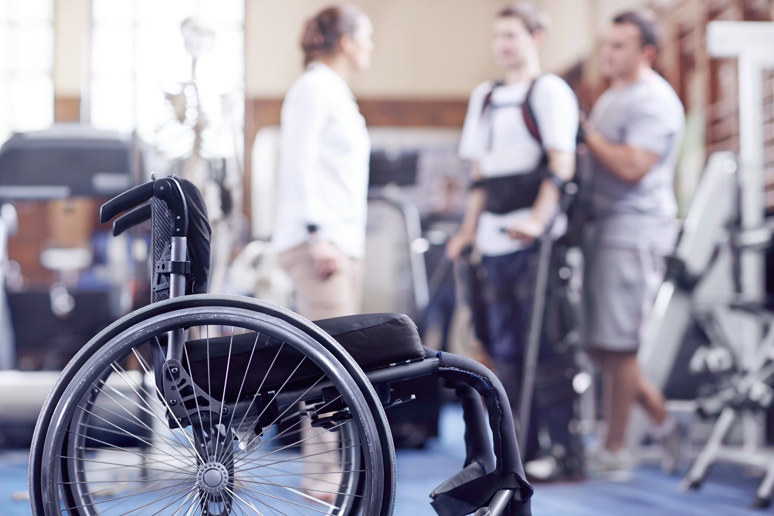 A close-up of an empty manual wheelchair sits in a rehabilitation clinic while a patient and medical staff stand blurred in the background, illustrating the long-term care needs managed by Tulsa spinal cord injury lawyers.