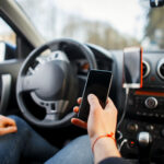 A close-up view of a driver's hand scrolling on a smartphone screen while behind the wheel of a car, demonstrating reckless actions that lead to distracted driving punitive damages OKC claims.