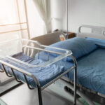 Empty hospital bassinet beside a delivery room bed.
