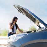 A stressed woman calling on her phone beside a car with an open hood illustrates what to do after a car accident in Tulsa.