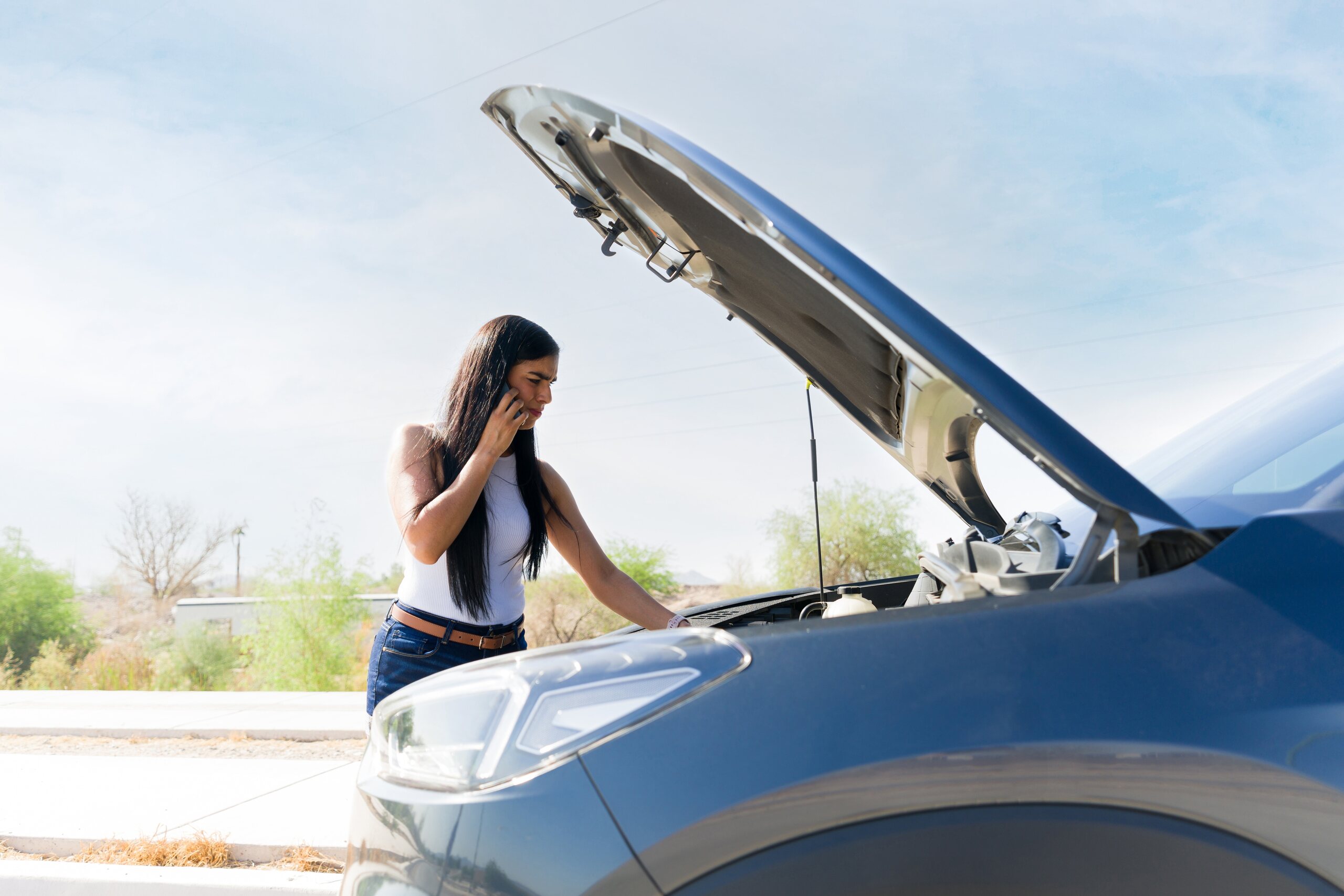 A stressed woman calling on her phone beside a car with an open hood illustrates what to do after a car accident in Tulsa.